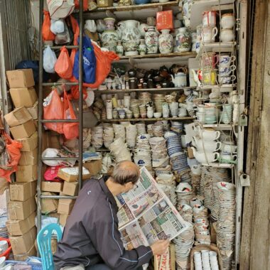 A merchant in an Asian ceramic shop surrounded by diverse pottery and plates.