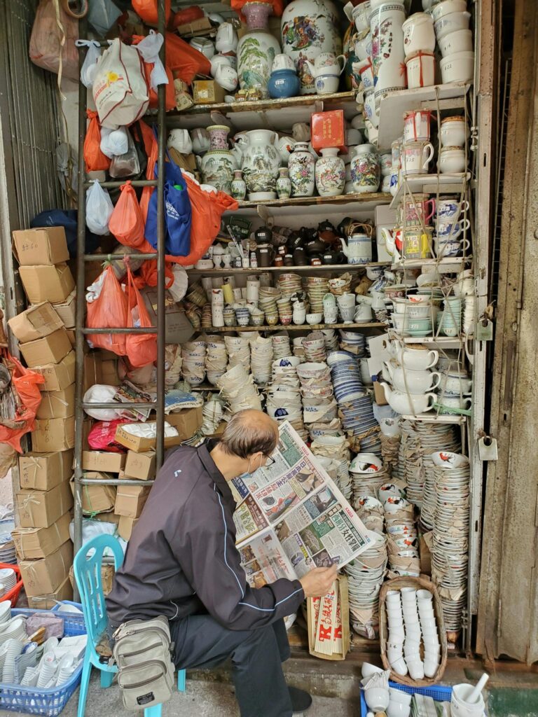 A merchant in an Asian ceramic shop surrounded by diverse pottery and plates.